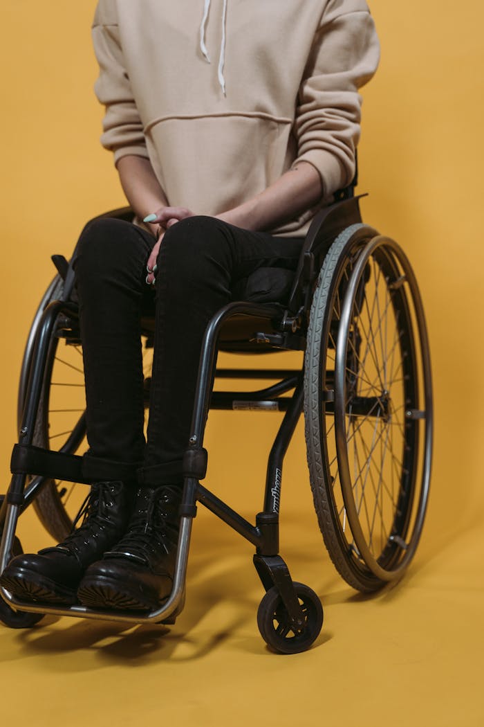 A person sitting in a wheelchair wearing black boots and a hoodie, set against a bright yellow backdrop.